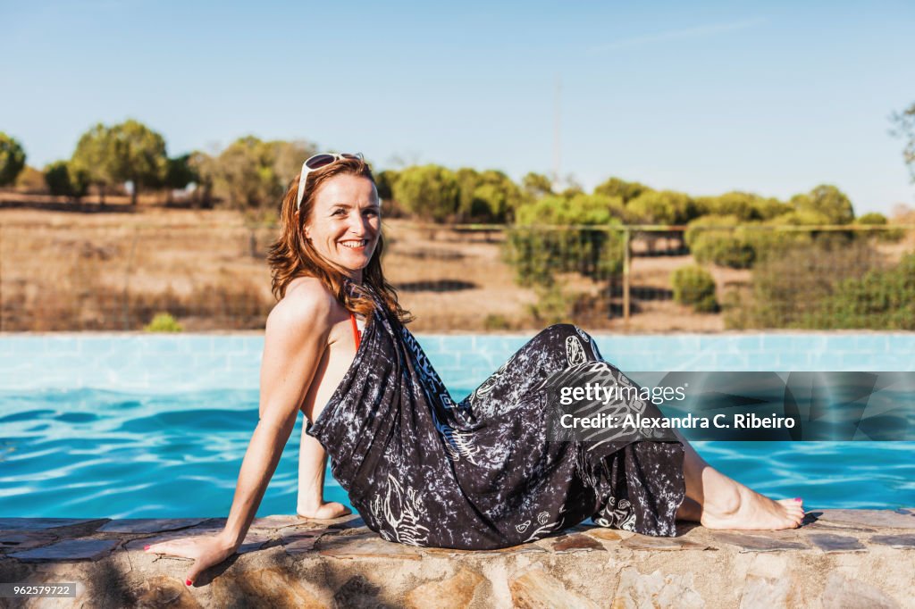 Side view portrait of smiling mid adult woman wearing sarong sitting at poolside on sunny day