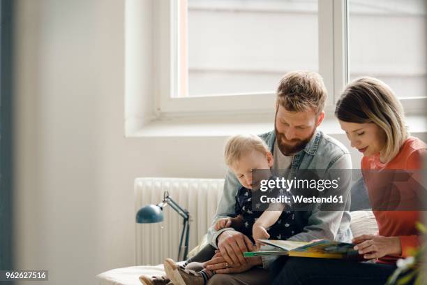 little boy sittiing on father's lap, mother reading out children's book - comfortable couple stock pictures, royalty-free photos & images