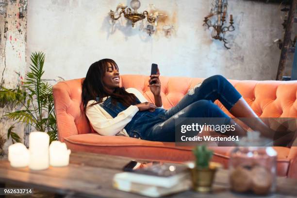laughing woman lying on the couch at home using cell phone - jeans latzhose frau stock-fotos und bilder