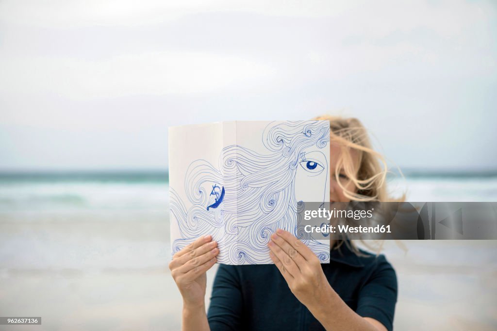Woman covering face with book, reading poetry on beach