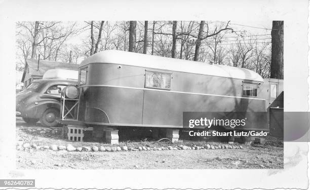 Black and white photograph, showing the side of a vintage caravan or towed trailer, resting on concrete blocks in a wintery landscape, with a...