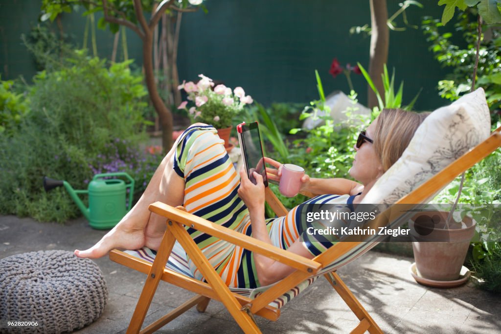 Woman relaxing on deck chair in backyard, reading on digital tablet