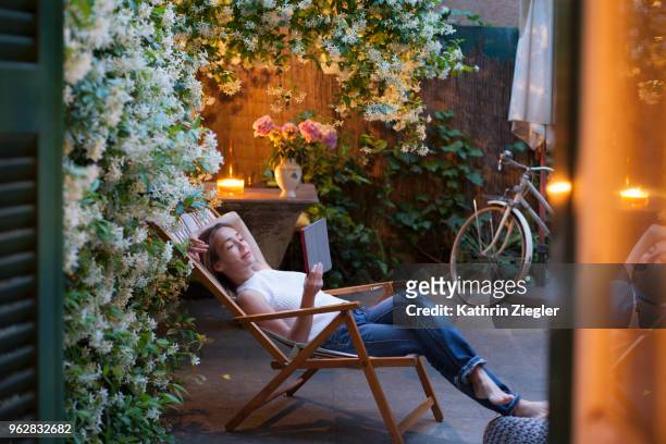 woman relaxing on deck chair in backyard at dusk, reading on digital tablet - adult woman garden flower stock-fotos und bilder