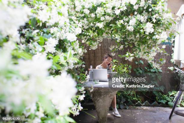woman sitting at garden table with laptop, enjoying her lush outdoors office - jasmin stockfoto's en -beelden