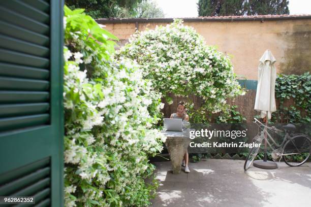 woman sitting at garden table with laptop, enjoying her lush outdoors office - kathrin ziegler stockfoto's en -beelden