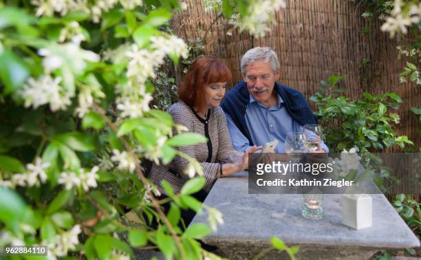 senior couple enjoying a glass of wine together at garden table, looking at pictures on smartphone - kathrin ziegler stockfoto's en -beelden