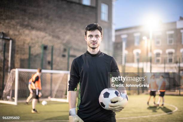 Waist up portrait of young man looking at camera and holding football in goalkeeper's kit