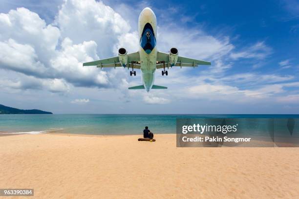 young man sitting on the beach and airplane in background at mai khao beach - aterrizar fotografías e imágenes de stock