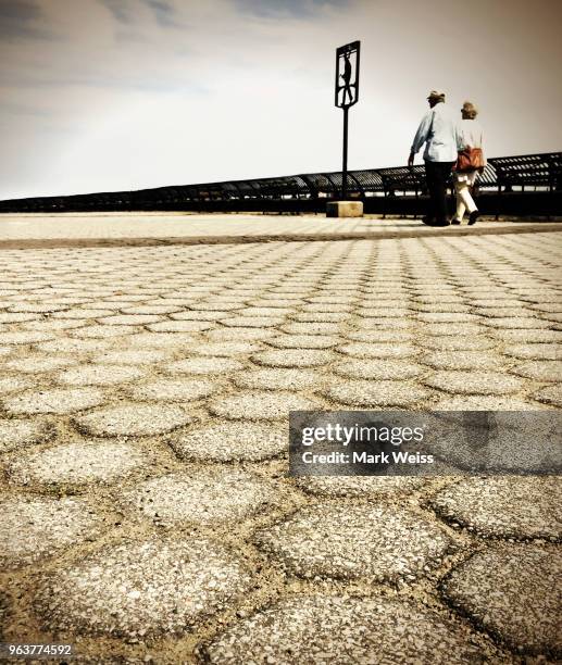 low angle view of mature couple strolling along iron railing on cobblestone path in new york with wispy overcast sky. desaturated color - limping stock pictures, royalty-free photos & images
