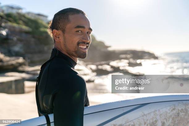 handsome surfer carrying his board and looking at the camera smiling - oceania stock pictures, royalty-free photos & images