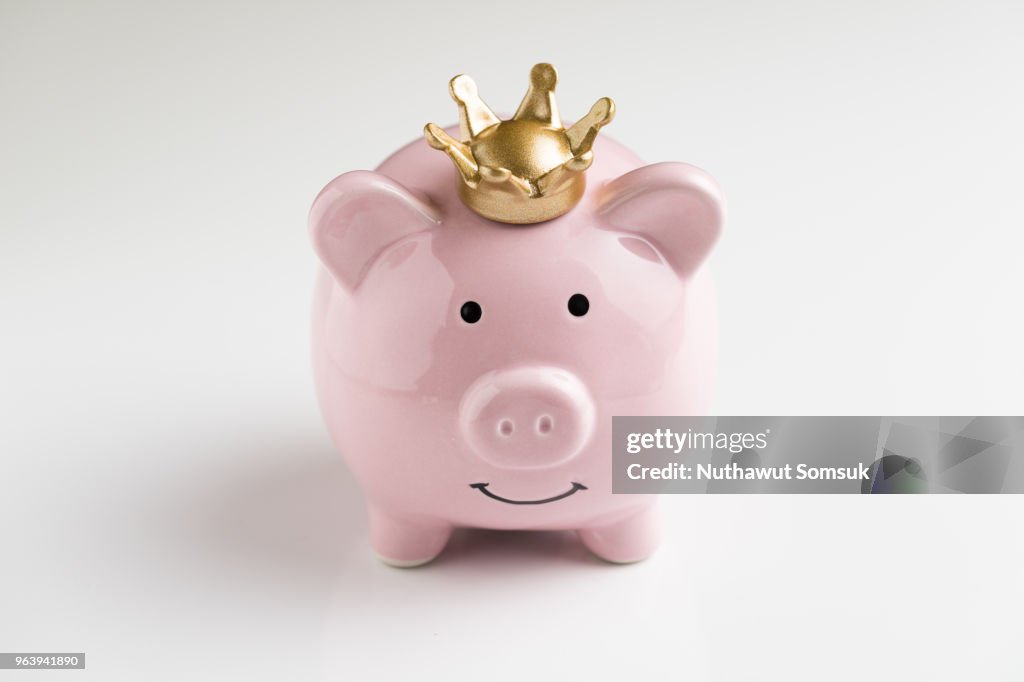 King of money savings concept, financial winner, smiling happy pink piggy bank wearing a golden crown on seamless white table background, bright future of compound interest investment