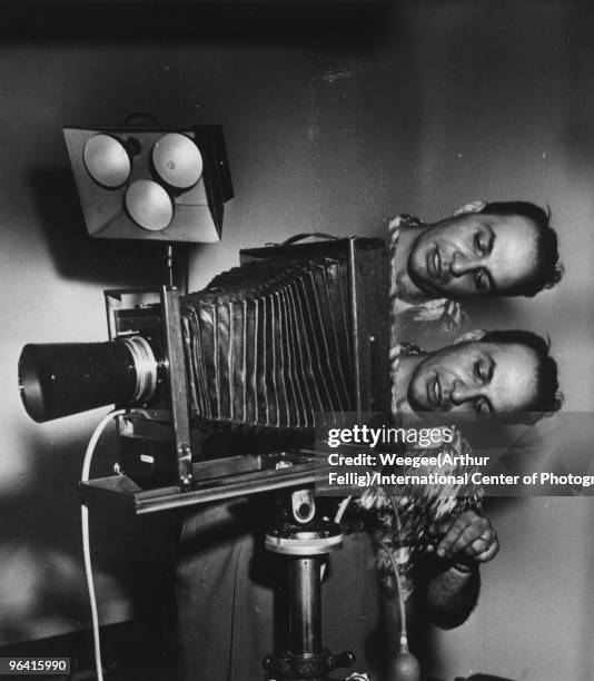 Distorted image of a man with two heads looking through the lens board of a large view camera on a tripod, with studio lights in the background, New...