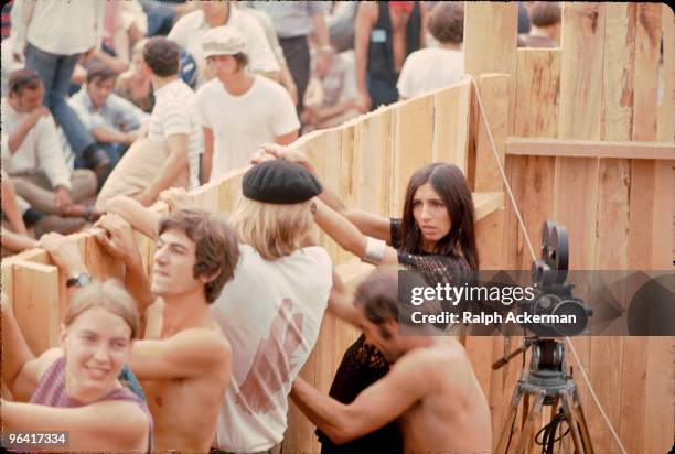 Near a tripod-mounted movie camera, a group of festival goers peer over the edge of a wooden fence during the Woodstock Music and Arts Fair, Bethel,...