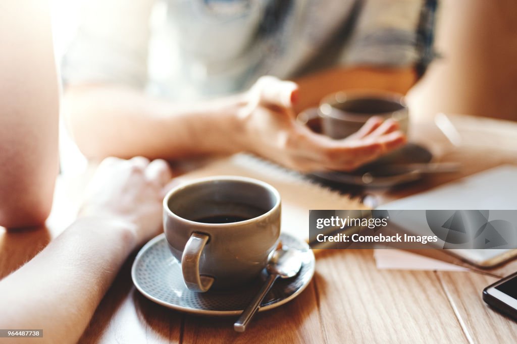 Friends talking at cafe table during coffee break. Unrecognizable male and female colleagues discussing business issues, focus on coffee cup with saucer and teaspoon