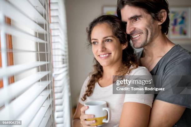 happy couple embracing next to window with a cup of coffee - jalousie stock pictures, royalty-free photos & images