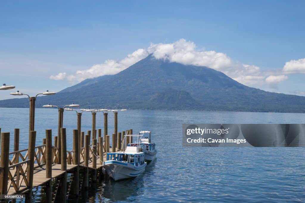 Boats moored by pier on Lake Atitlan against mountains and cloudy sky
