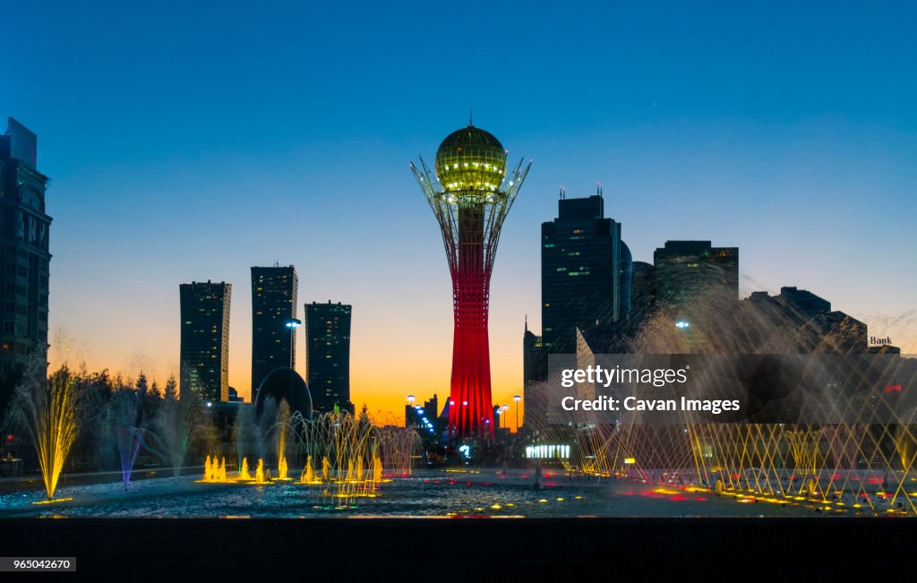 Illuminated Bayterek tower and fountain in city against clear blue sky