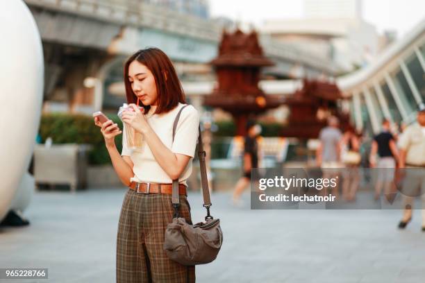 jeune femme asiatique dans le quartier du centre-ville de bangkok, prendre une pause, envoyer des sms sur la cellule tout en sirotant une boisson - sms'en photos et images de collection