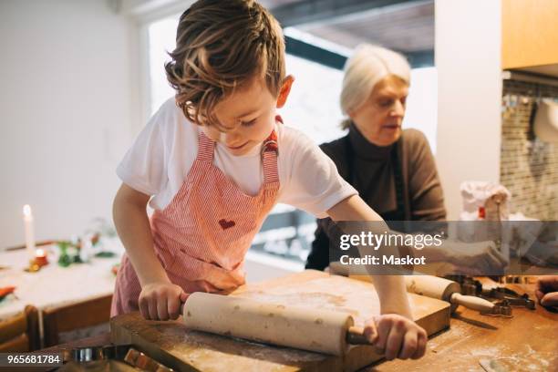 boy rolling dough while standing by grandmother at kitchen counter - generationen kochen stock-fotos und bilder