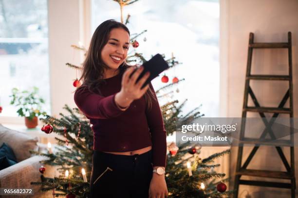 smiling woman taking selfie through smart phone against christmas tree at home - national day in sweden 2017 stock pictures, royalty-free photos & images