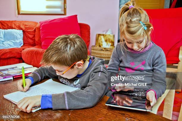 Petit garcon de 9 ans écrivant sur un cahier et petite fille de 6 ans jouant sur sa tablette numérique.