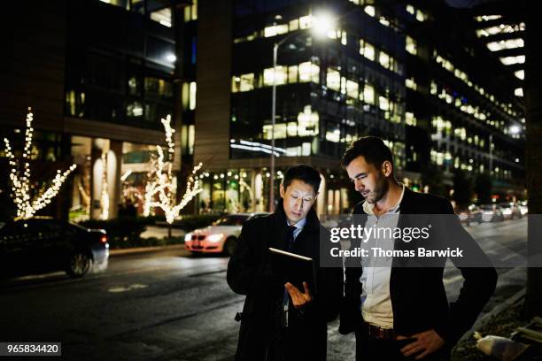 Two businessmen standing on city street at night while looking at data on digital tablet