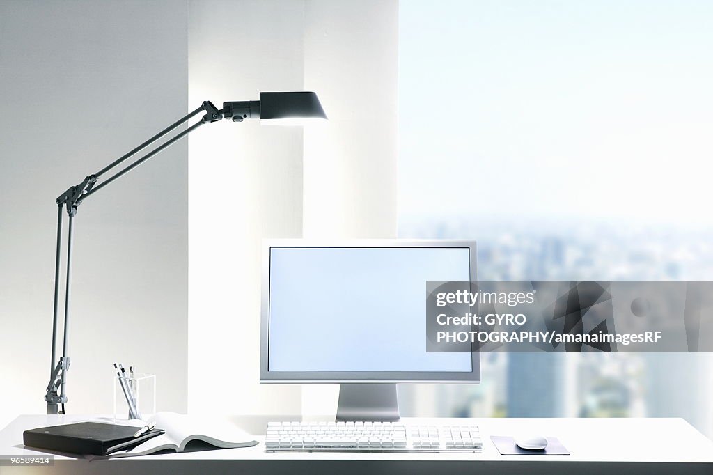 Desk lamp and computer on desk with view of a city through the window