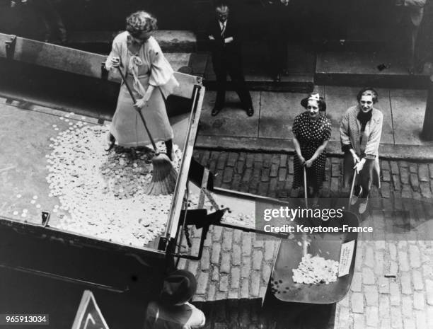 Une femme, debout sur un camion rempli de pièces d'argent, pousse les pièces avec un balai pour les faire tomber dans une brouette et ensuite les...