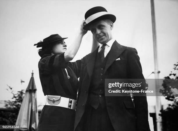 Une femme portant une ceinture blanche sur laquelle est accroché un porte-monnaie, attache sur le chapeau melon d'un homme un ruban blanc, à Londres,...