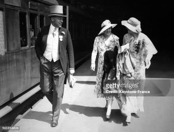 Un homme en redingote et chapeau haut-de-forme en compagnie de deux femmes portant des chapeaux et des robes élégantes sur le quai de la gare de...
