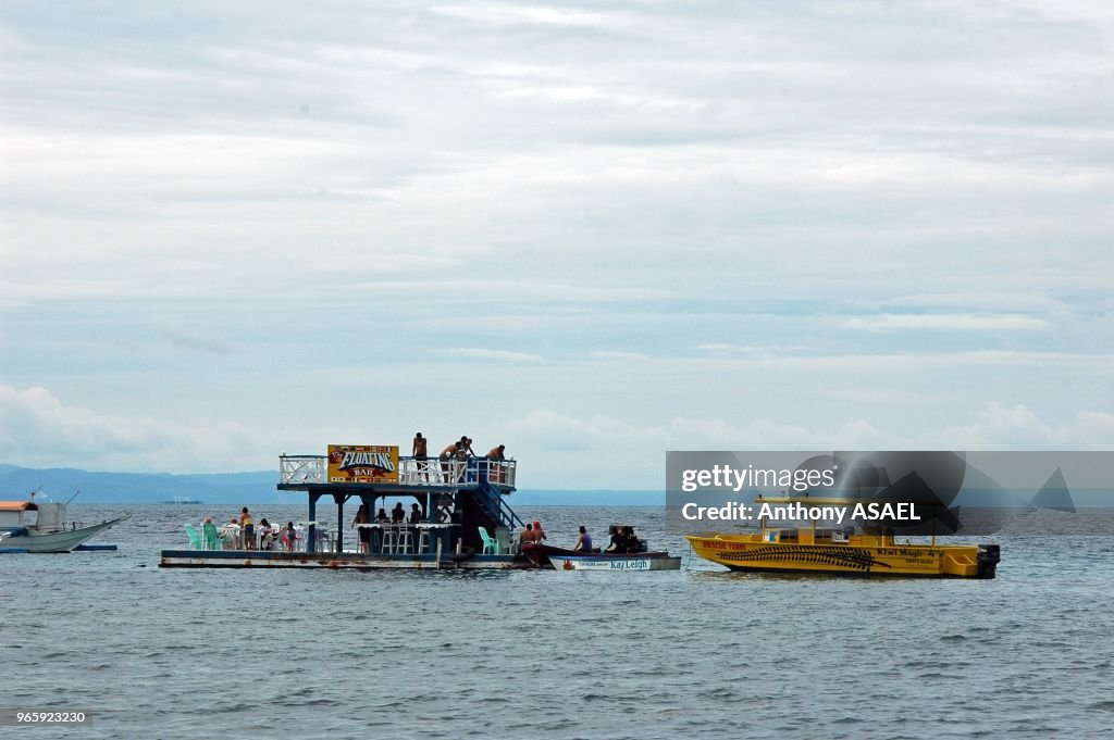 Coco Beach, Puerto Galera, Mindoro Island, Philippines, platform floating on the sea water with a small house used as a bar on it, some tourists sitting on chair on the terrasse, the mainland and its mountains, hills and volcanoes in the background.