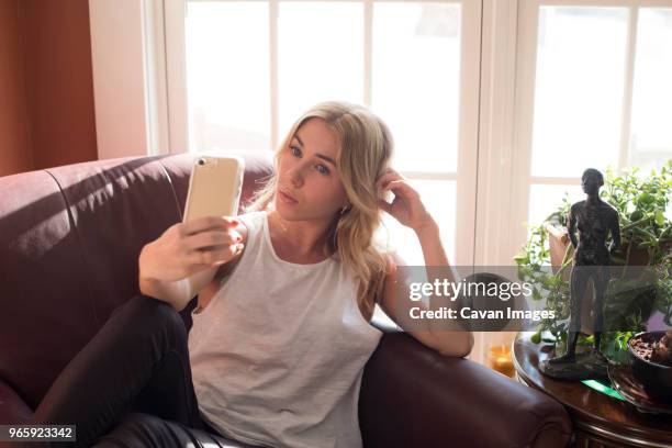 woman taking selfie while sitting on armchair at home - mobile sculpture fotografías e imágenes de stock