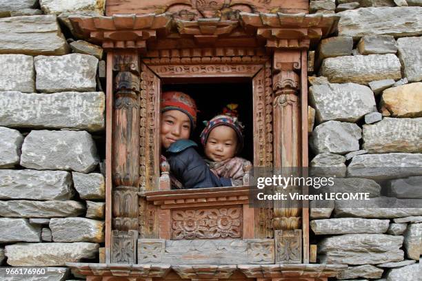 Deux enfants népalais regardent par la fenêtre de leur maison traditionnelle en bois sculpté caractéristique de l?artisanat Tamang dans le village de...