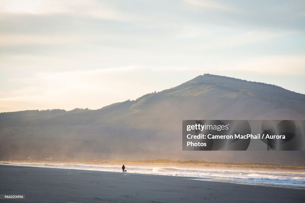 Woman walking dog at sunrise on beach of Netarts Bay, Oregon, USA
