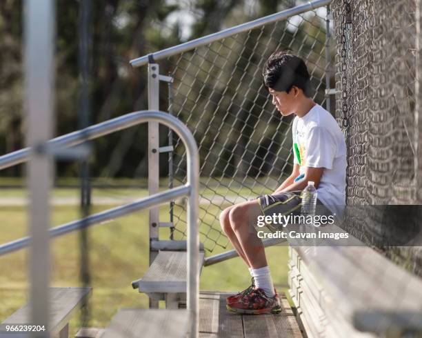 thoughtful teenage boy sitting on bench against chainlink fence at playing field - chainlink fence stock pictures, royalty-free photos & images