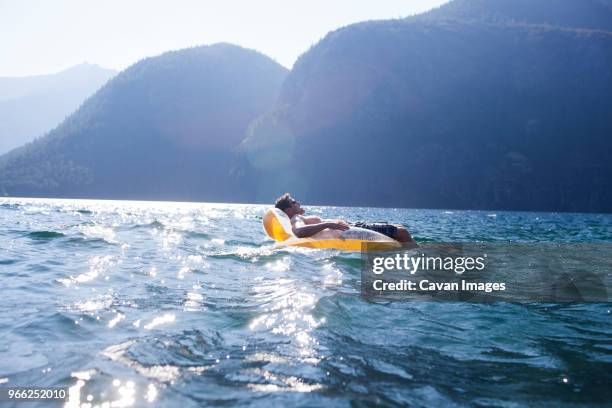 side view of man lying on pool raft in lake against mountains during summer - luftmatraze stock-fotos und bilder