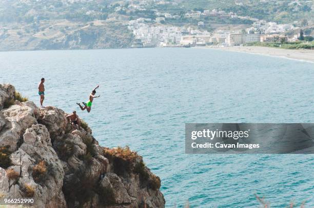 friends looking at man jumping into sea from cliff - cliff jumping stock pictures, royalty-free photos & images