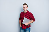 Portrait of smart clever qualified promising it-specialist software developer dressed in red checkered casual shirt, jeans, holding closed netbook in hands, isolated on gray background