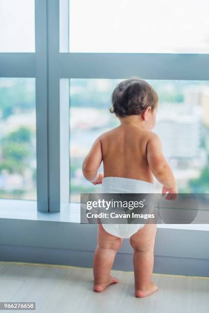 baby boy looking through window while standing at home - diaper boy fotografías e imágenes de stock