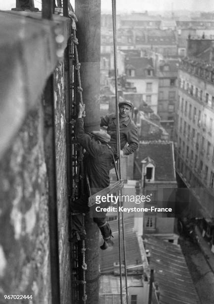 Suspendus à des cordes instables, ces ouvriers fixent sur un toit de la Cité Berryuer une cheminée métallique, à Paris, France le 21 octobre 1932.