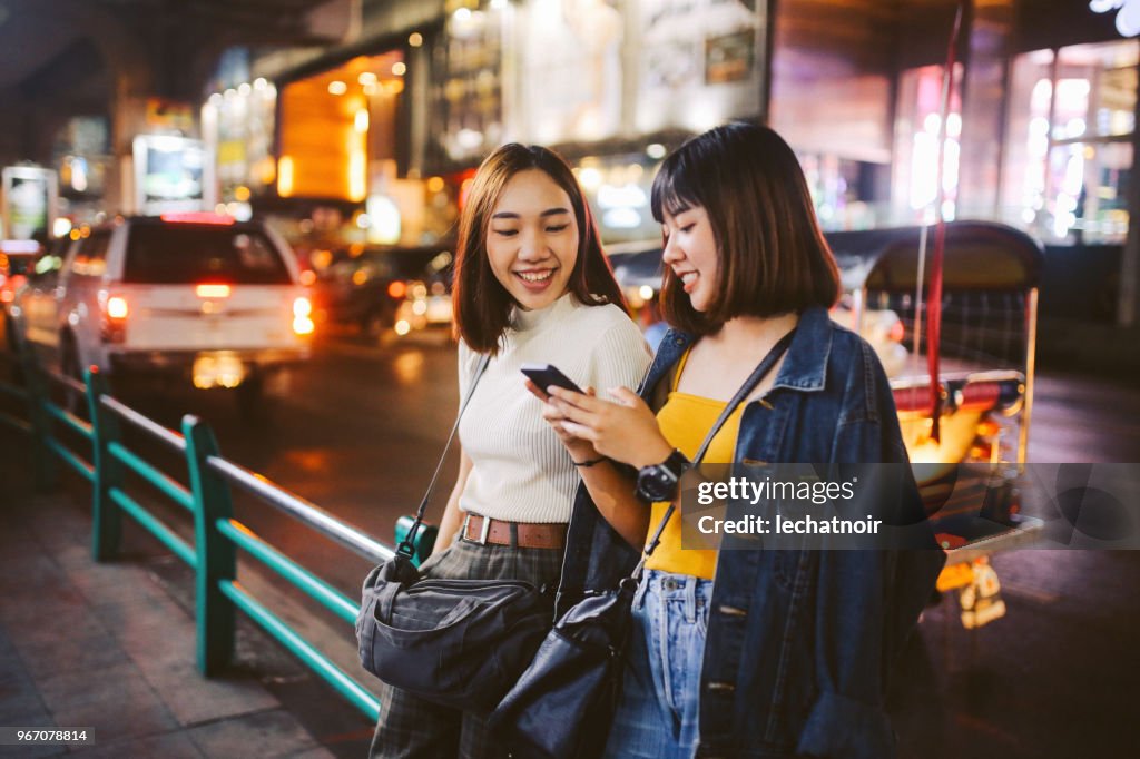 Dos chicas asiáticas en Bangkok, buscando en sus smartphones, charlando sobre los medios de comunicación social