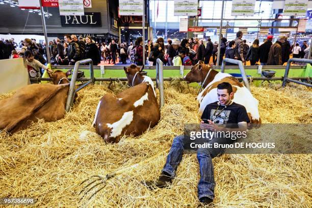 Un jeune homme allongé à côté d'une vache et lisant son smartphone lors du Salon International de l'Agriculture le 25 Février 2017 à Paris, France.