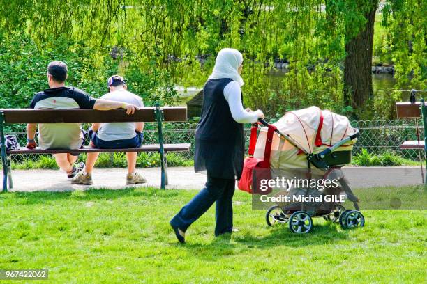 Femme avec foulard sur sa tete roule une poussette sur la pelouse du Jardin des Plantes, Le Mans, France.