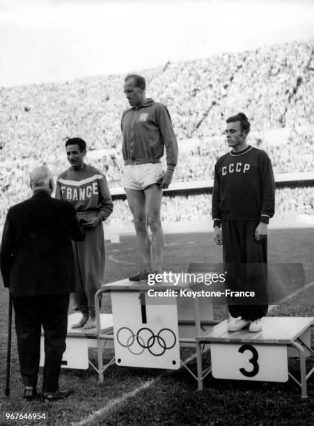 Sigfrid Edstrom, président du comité olympique, remet la médaille d'argent au coureur français Alain Mimoun qui se tient sur le podium à côté du...