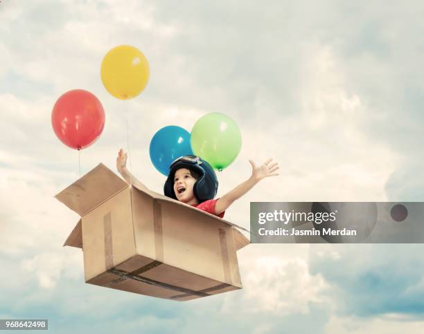 kid flying in cardboard box with balloons between clouds - boy in a box stock-fotos und bilder