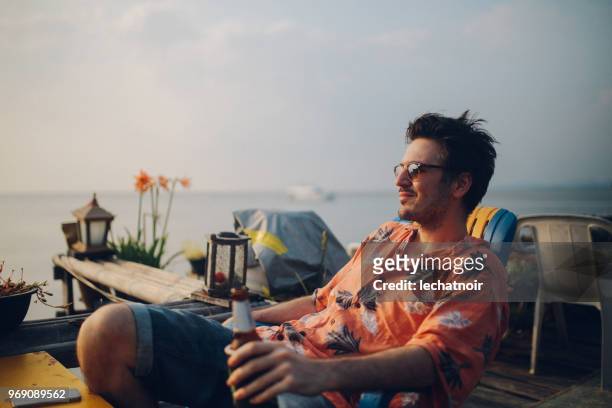 young man relaxing by the ocean, having a beer, looking at sunset at koh lanta, thailand - hawaiian shirt imagens e fotografias de stock