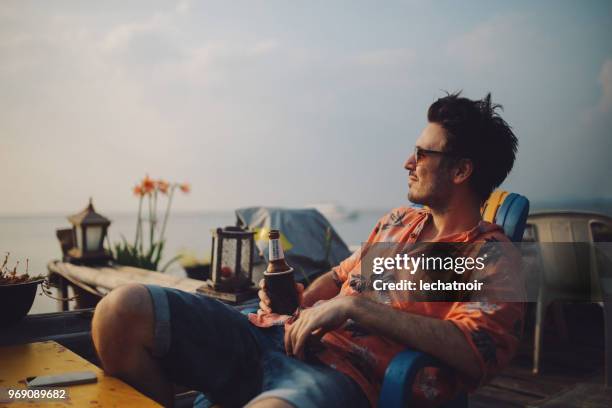 junger mann am meer entspannen, ein bier, blick auf sonnenuntergang auf koh lanta, thailand - hawaiian shirt stock-fotos und bilder