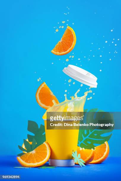 orange juice dynamic splash in a paper cup with flying orange slices and tropical leaves, action food photography on a bright blue background with copy space. summer refreshing drink. - orange juice glass white background stock pictures, royalty-free photos & images