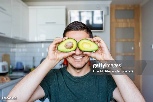 young man having fun with avocado at the kitchen - funny vegetable stockfoto's en -beelden