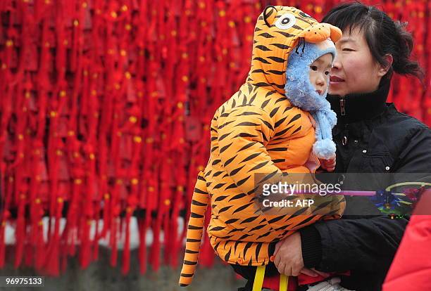 Woman carries a child dressed in a tiger outfit past lucky votives hung on a railing on the first day of the Chinese New Year at Dongyue Miao, the...